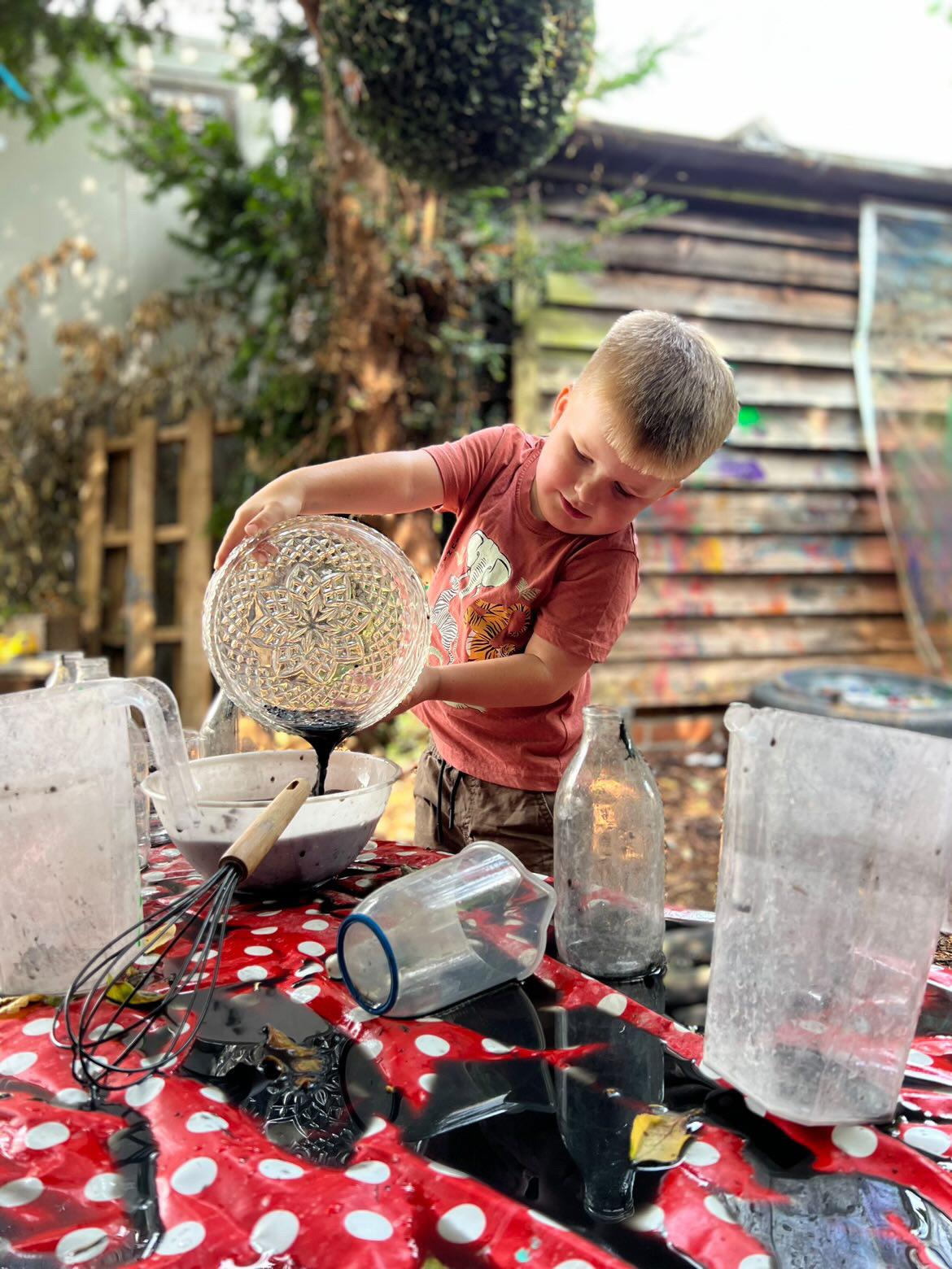 A boy pouring a dark potion from a glass bowl at the mud kitchen