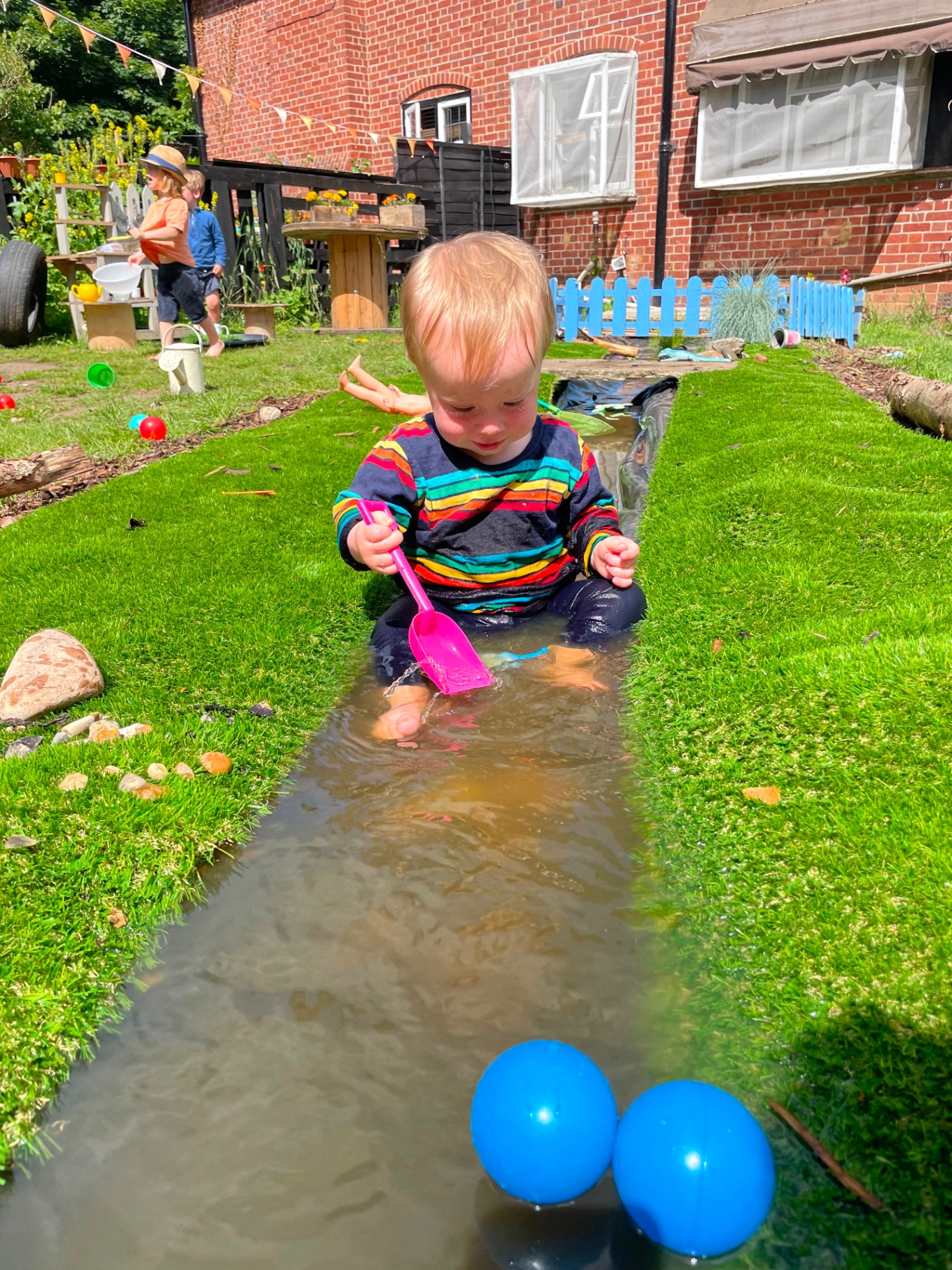 A toddler sitting in a shallow stream with a spade