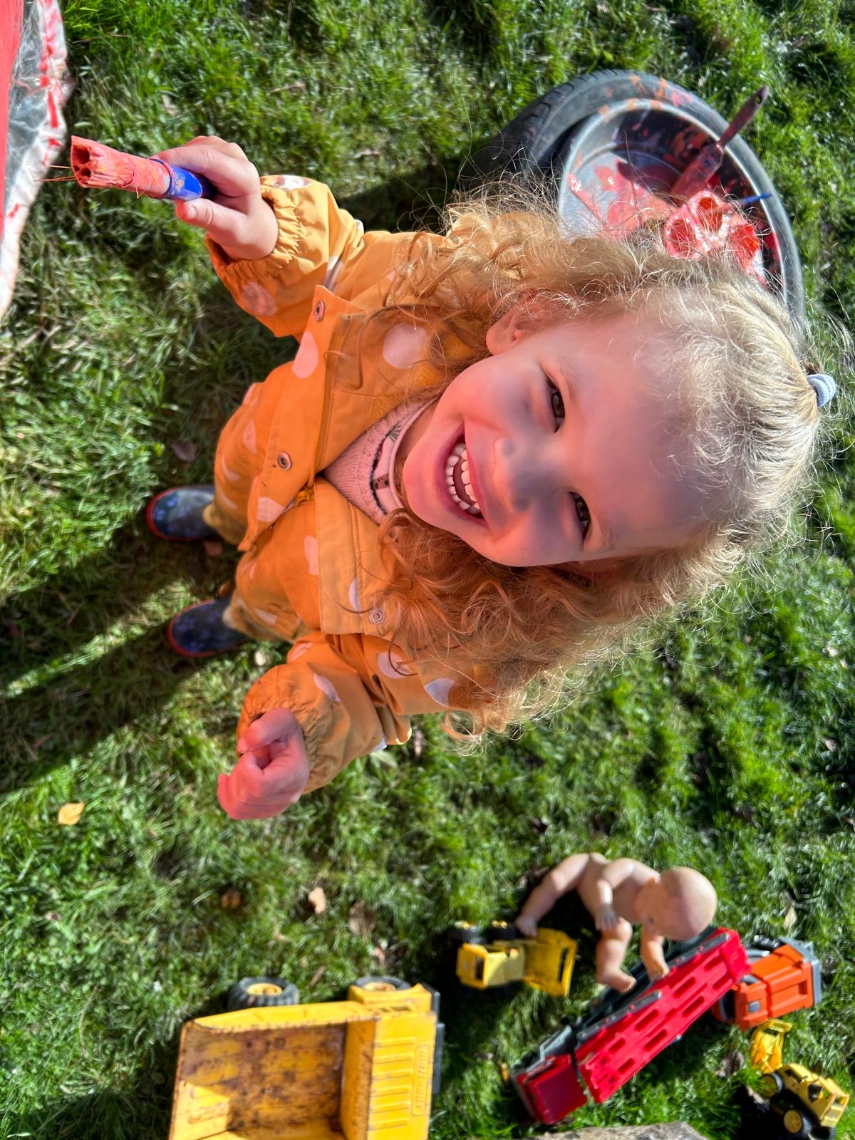 A grinning child holding up a paintbrush in the sunshine