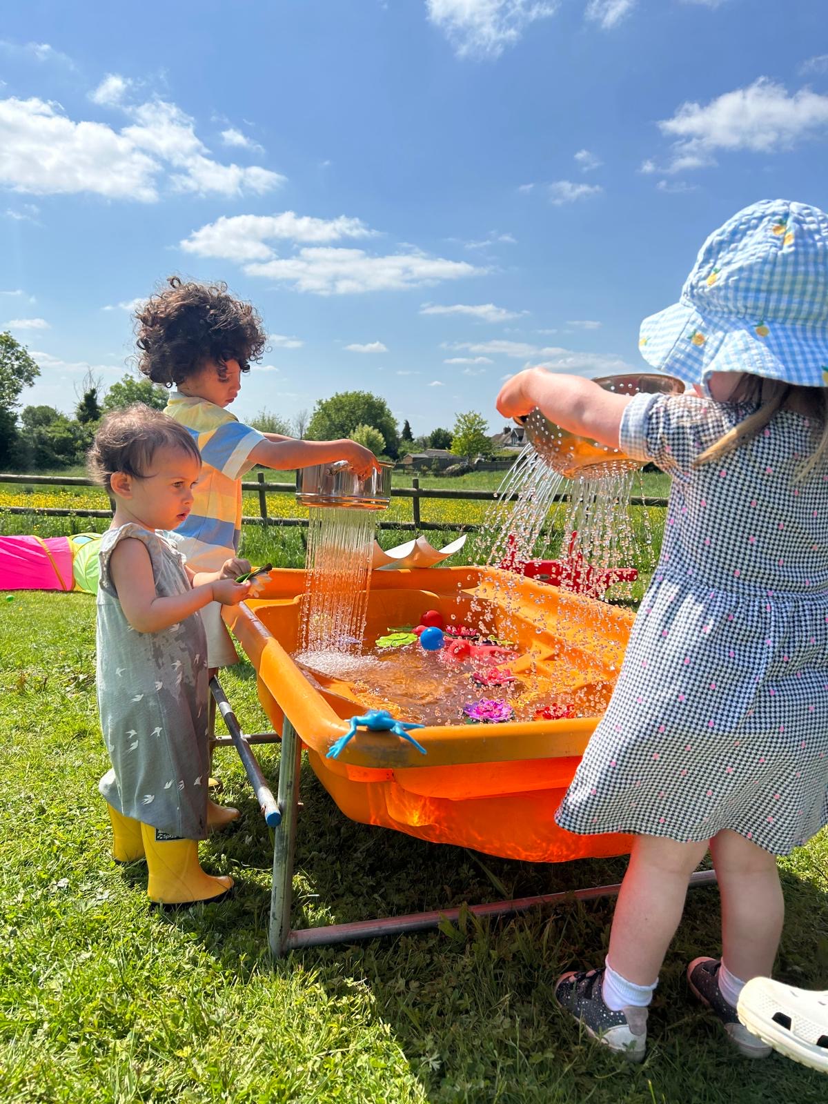 Children pouring water at an orange water table under a blue sky