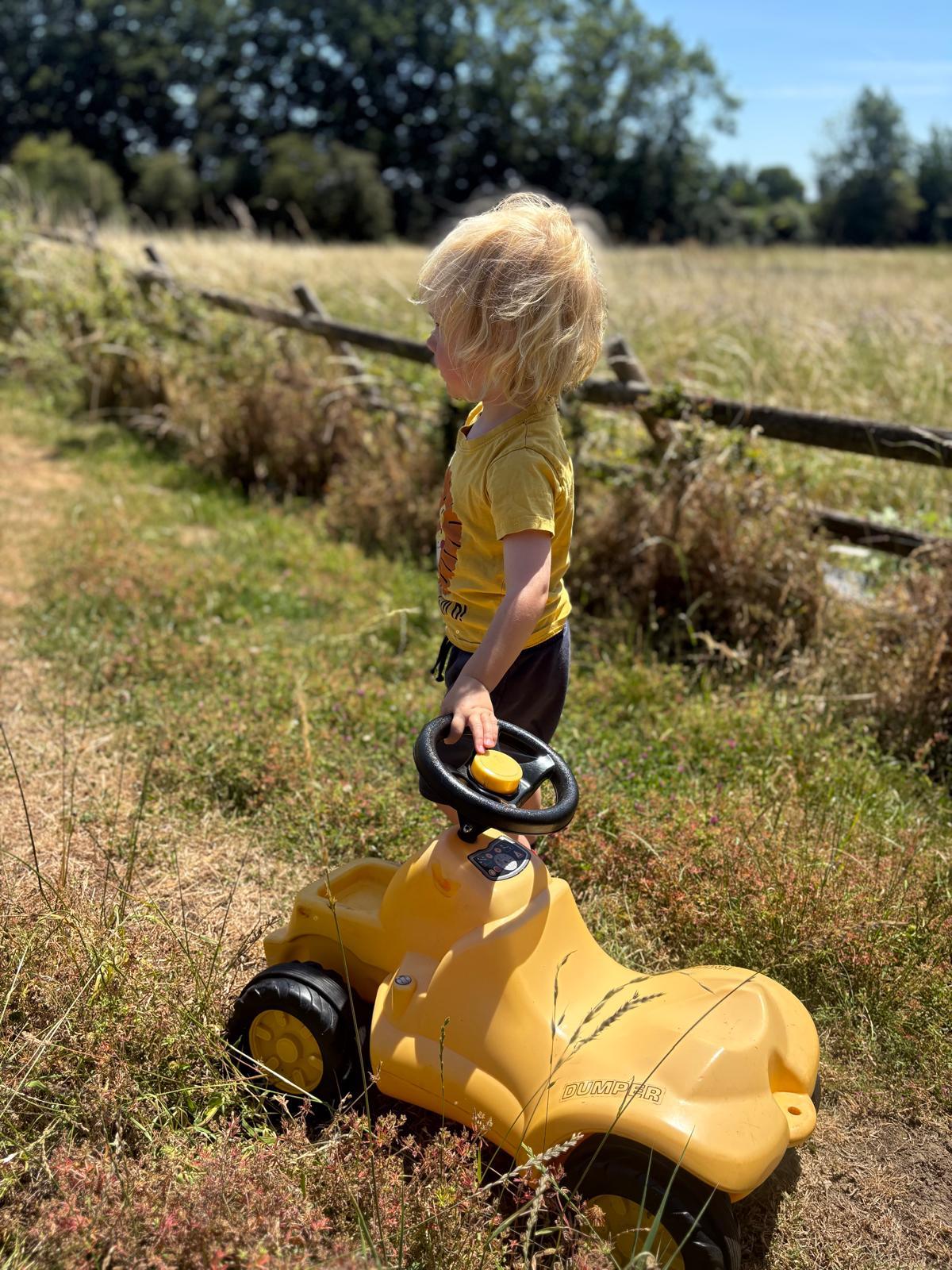 A child pushing a yellow ride-on dumper truck through a sunny meadow