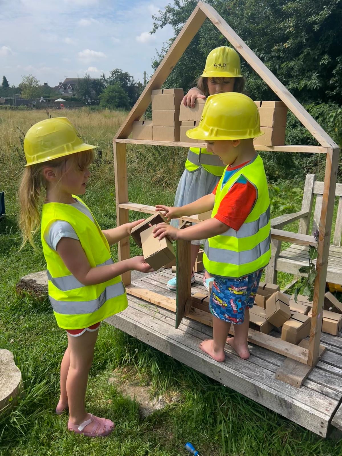 Three children in hard hats and hi-vis building with cardboard bricks