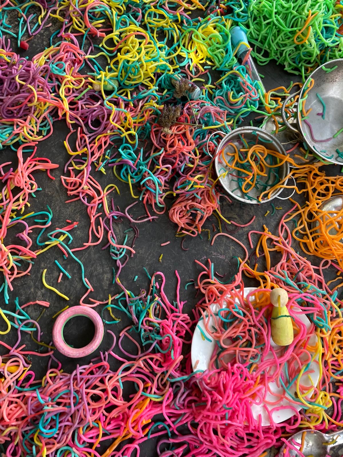 Rainbow coloured spaghetti spread across a tuff tray with tin pots